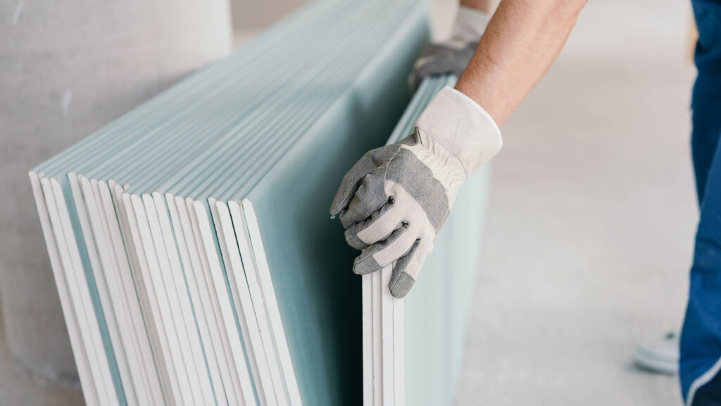 Ein Stapel Gipskartonplatten lehnt an der Wand. Großaufnahme auf die Hände eines Handwerkers in weiß-grauen Handschuhen, der vier der Platten wegnimmt.