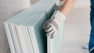 Ein Stapel Gipskartonplatten lehnt an der Wand. Großaufnahme auf die Hände eines Handwerkers in weiß-grauen Handschuhen, der vier der Platten wegnimmt.