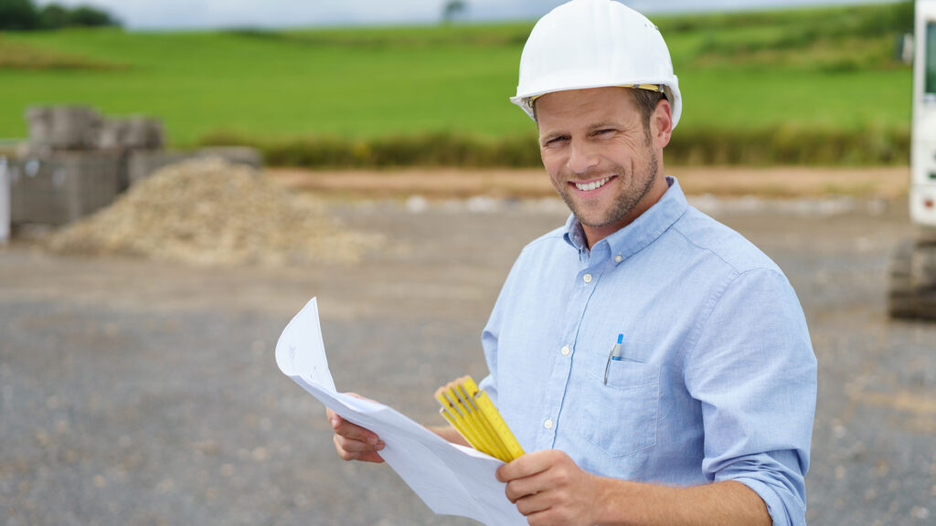 Ein Architekt steht mit Bauplan und Zollstock in der Hand an der Baustelle